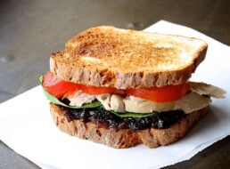 a white plate with a toasted chicken sandwich topped with California Prune bacon jam, tomatoes and lettuce