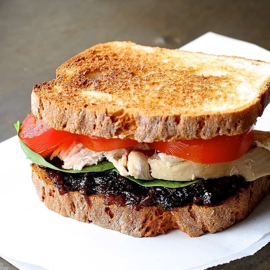 a white plate with a toasted chicken sandwich topped with California Prune bacon jam, tomatoes and lettuce