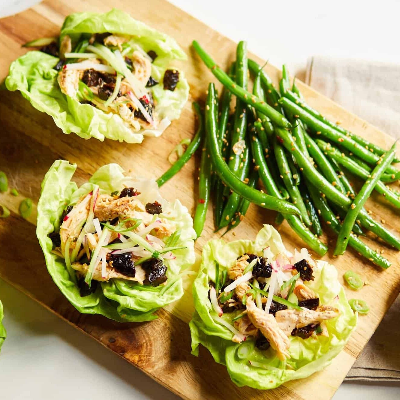 Three Asian-Inspired chicken lettuce wraps on a board with sesame green beans and prunes