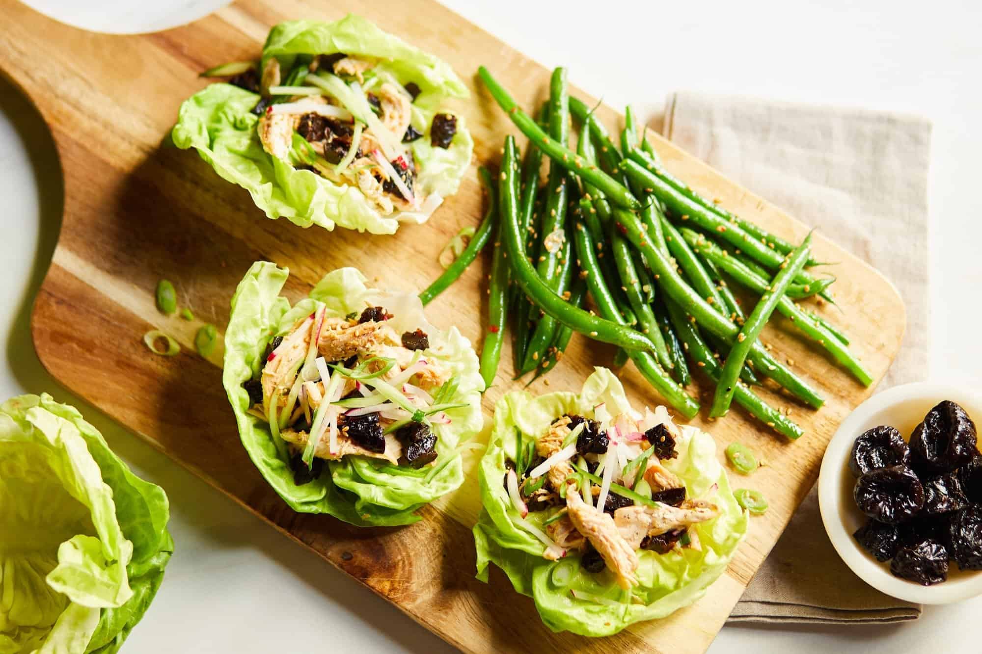 Three Asian-Inspired chicken lettuce wraps on a board with sesame green beans and prunes