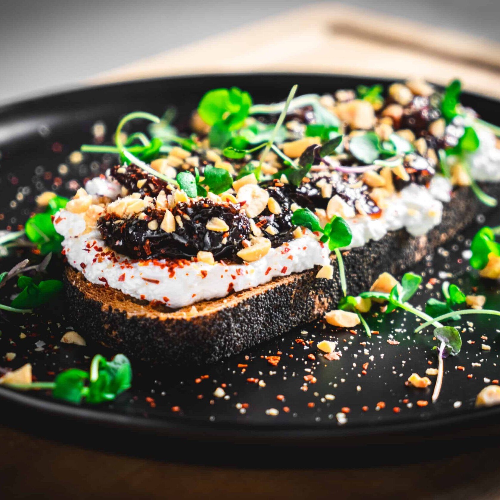 black plate with a piece of toast covered with ricotta, prune jam, peanuts and microgreens