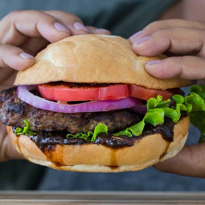 Two hands holding a burger stacked with prune sauce, onions, lettuce and tomatoes