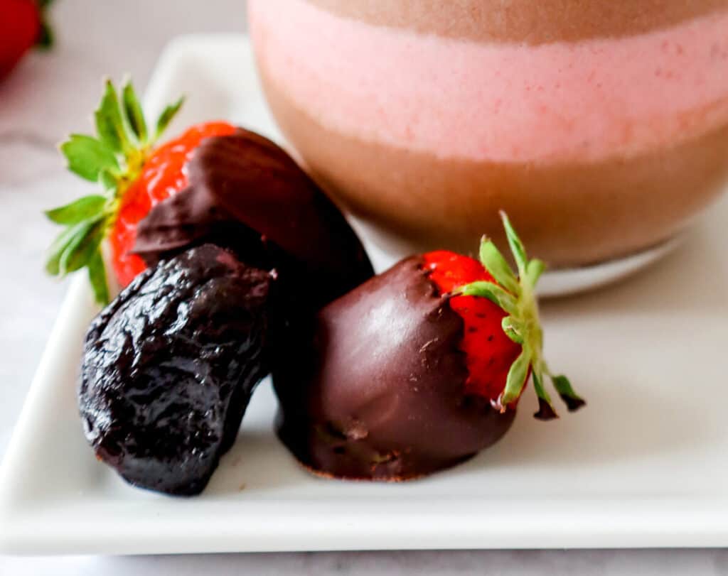 Two chocolate covered strawberries and a prune with a chocolate and strawberry layered smoothie in the background