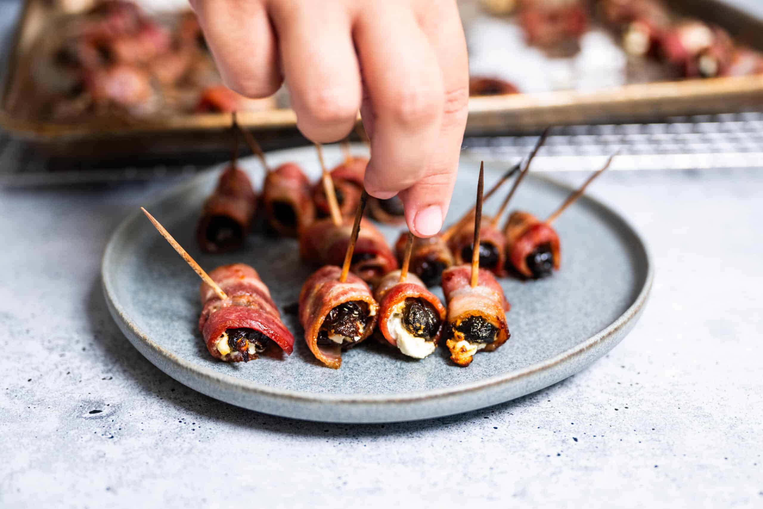 Devils on Horseback - Prunes stuffed with Goat cheese and wrapped in bacon Hand reaching to grab a bacon wrapped prune