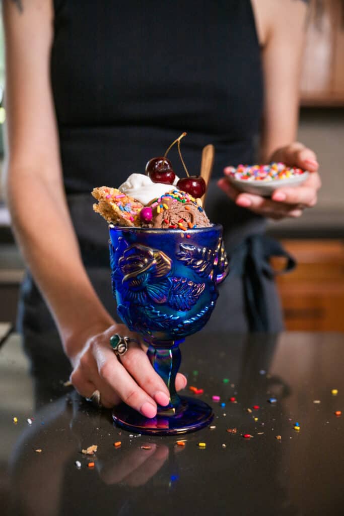 hand holding a blue glass filled with Vegan Chocolate Ice Cream (Nice Cream) Sundaes with whipped cream, sprinkles and a cherry