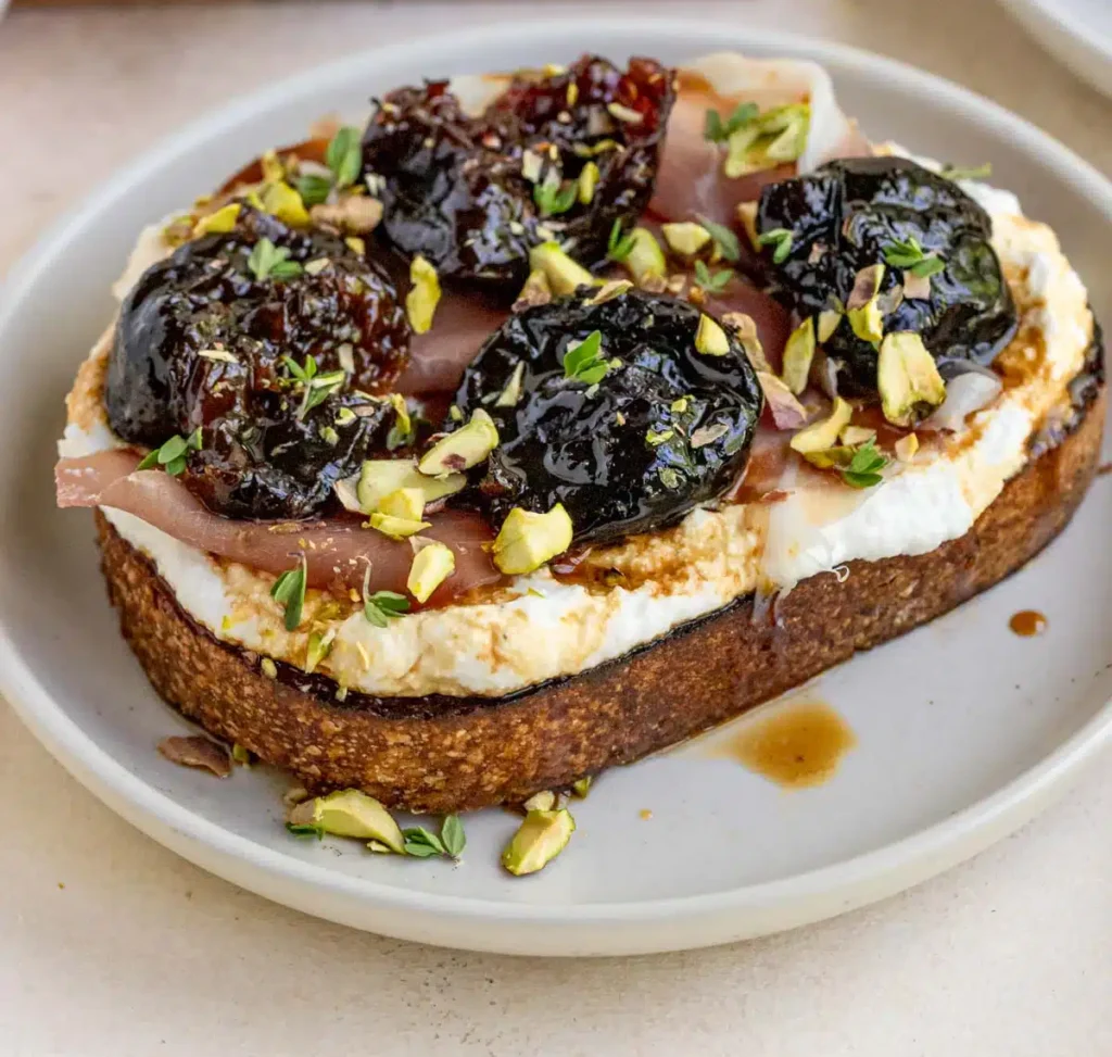 a slice of toast with ricotta, prosciutto and prunes sprinkled with pistachios