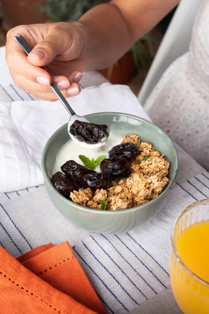 Person holding a spoon digging into a bowl of yogurt topped with whole prunes and granola.
