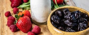 Bowl of prunes on cutting board with fresh strawberries and raspberries, spinach and a glass of milk