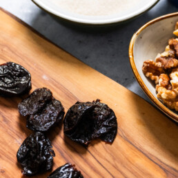 Prunes scattered on a cutting board with bowls of walnuts and seasonings placed off to the side