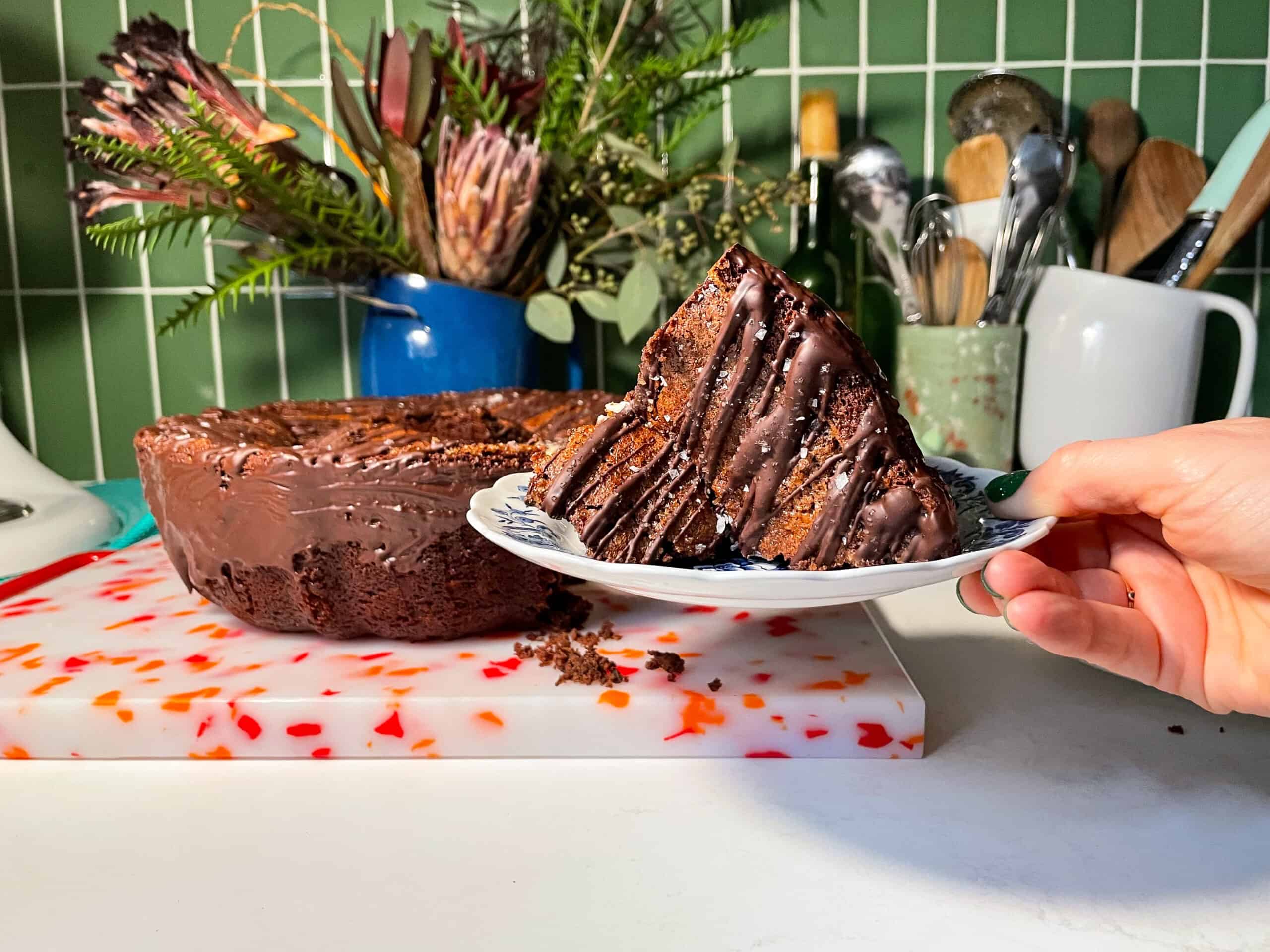 Hand holding a plate with a slice of chocolate cake. Remainder of cake plated on cutting board in kitchen appears in the background.