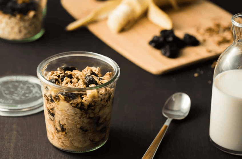 Mason jar of overnight oats on table with spoon, glass of milk and cutting board with prunes, nuts and a banana on top