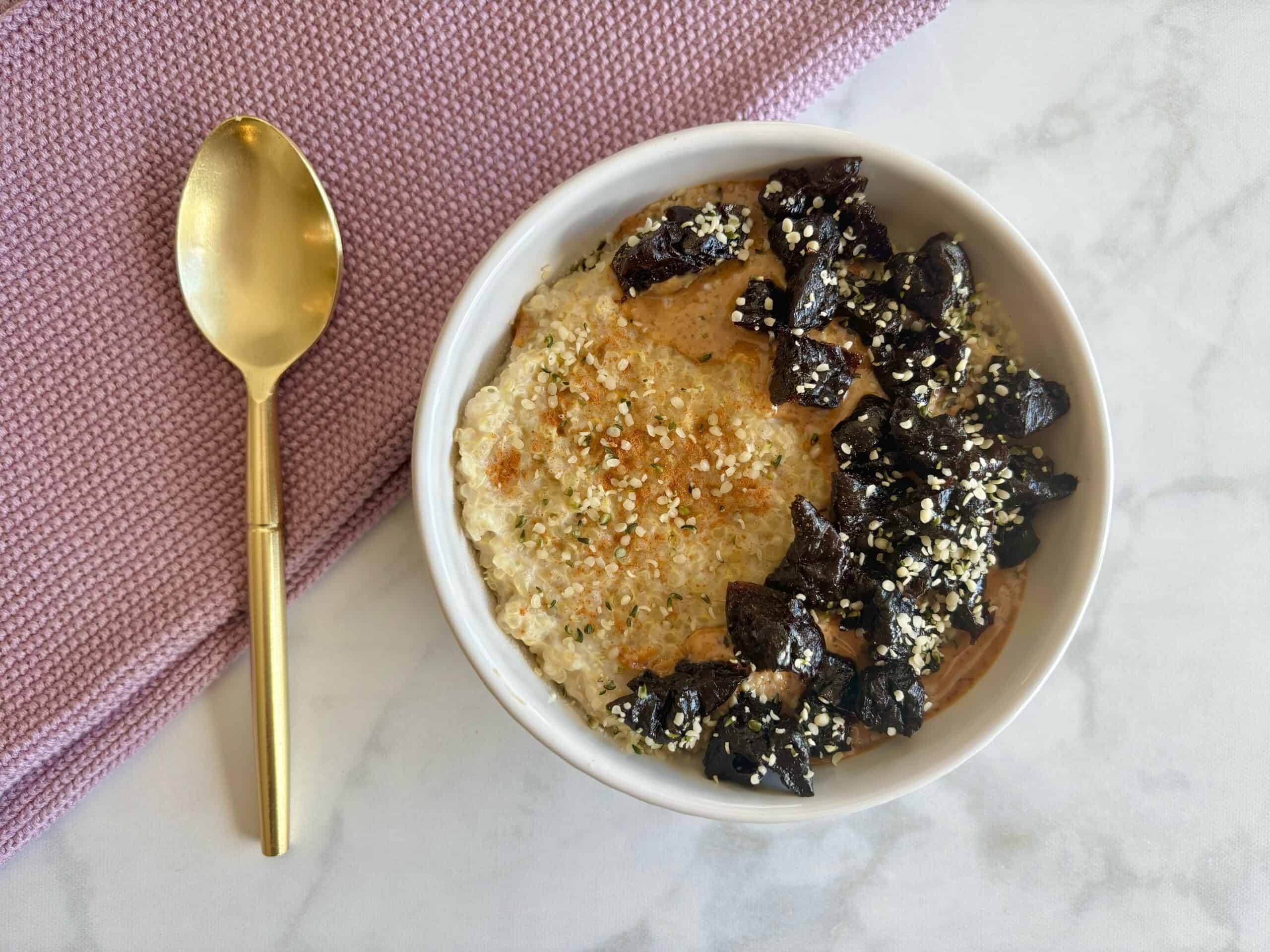 Bowl of porridge topped with prunes and hemp seeds with a golden spoon and purple dish towel underneath