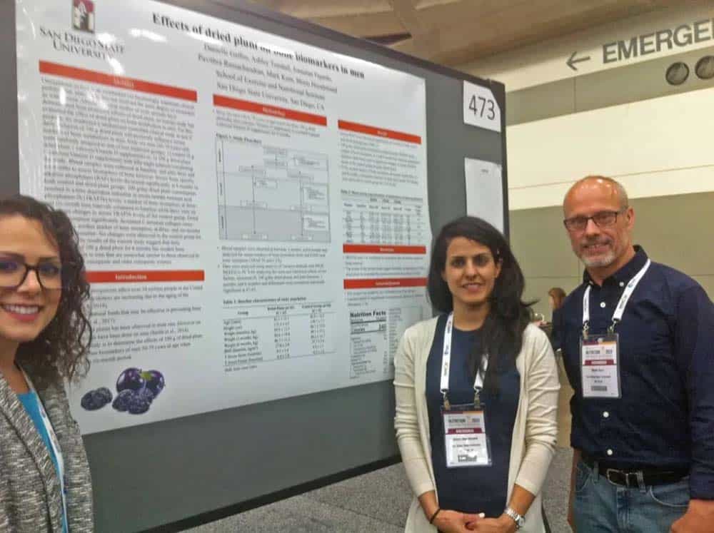 Two women and a man standing in front of a bulletin board with information about prunes and biomarkers in men.