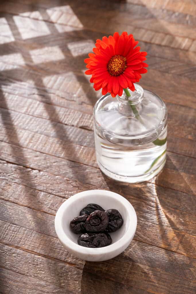 prunes in a white bowl next to a red flower in a vase