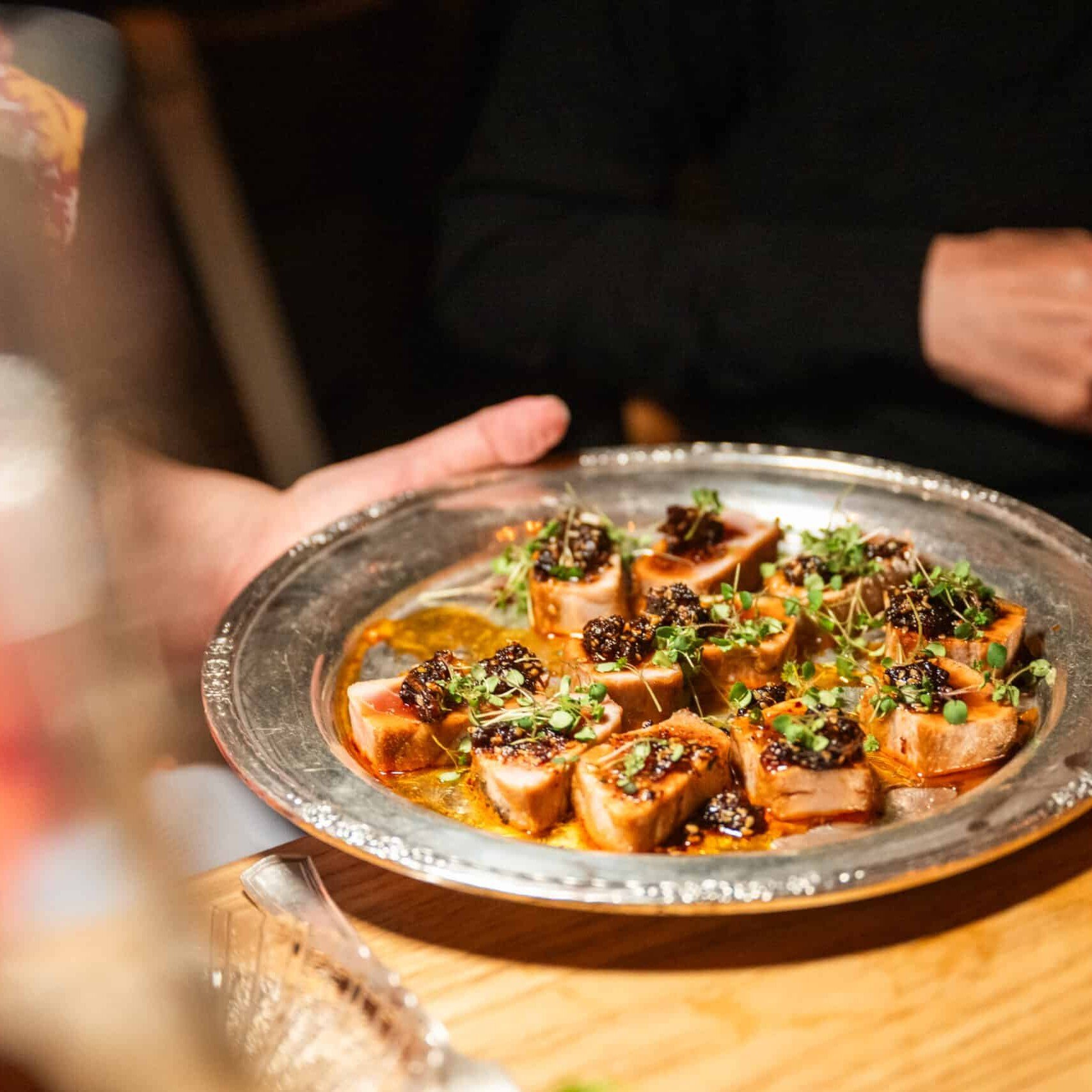 slices of grilled fish on a silver platter topped with pickled prunes