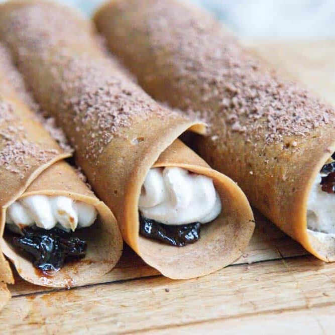 Four coffee soaked prune and whipped ricotta filled crepes on a serving board.