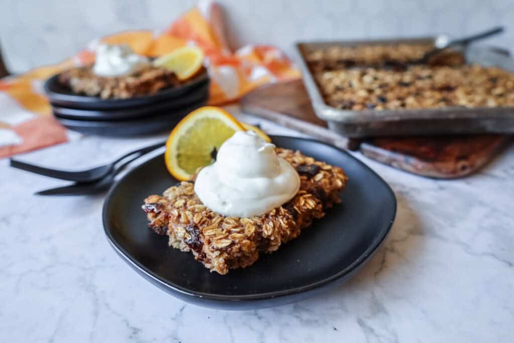 a slice of sheet pan baked oatmeal topped with yogurt and a sliced orange on a black plate