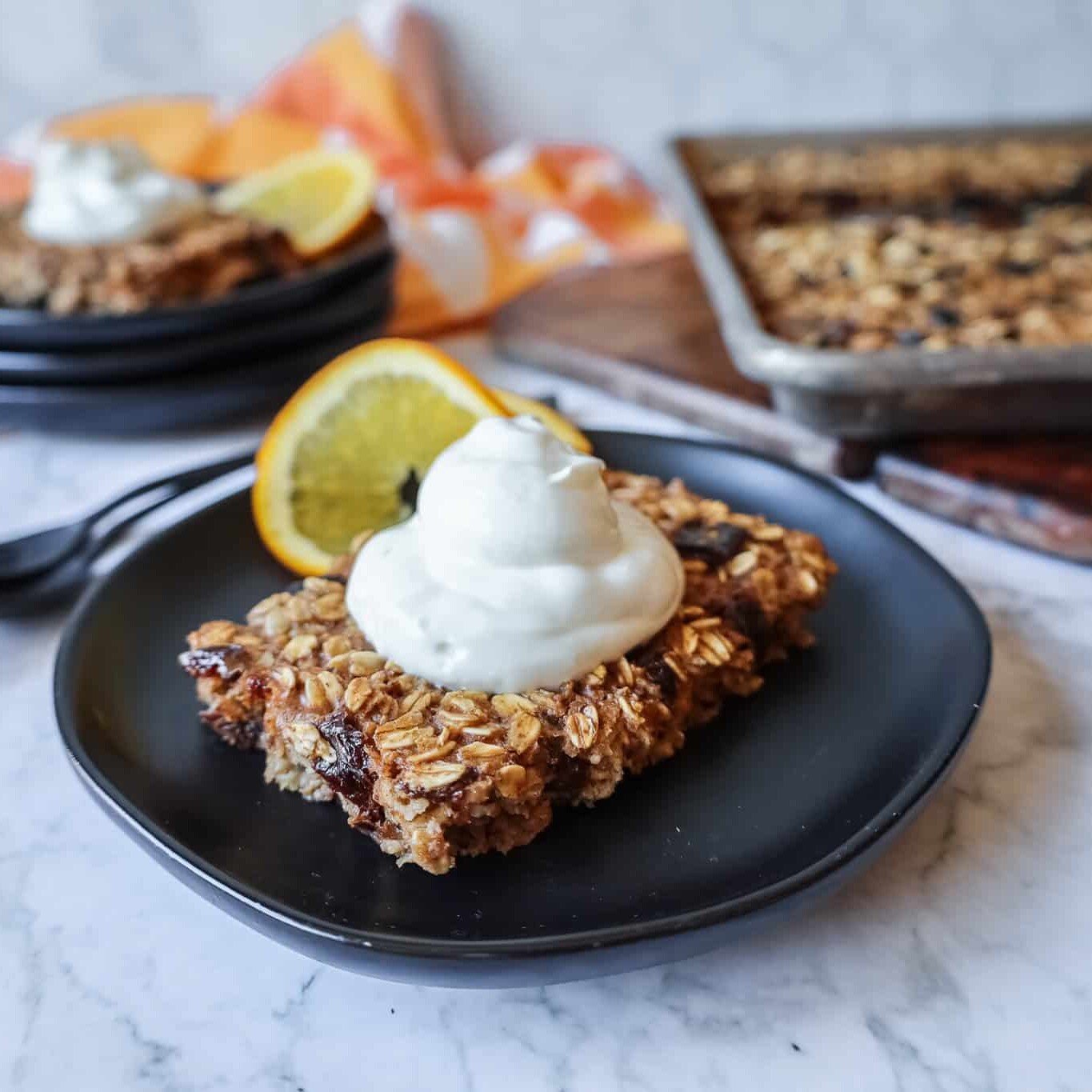 a slice of sheet pan baked oatmeal topped with yogurt and a sliced orange on a black plate