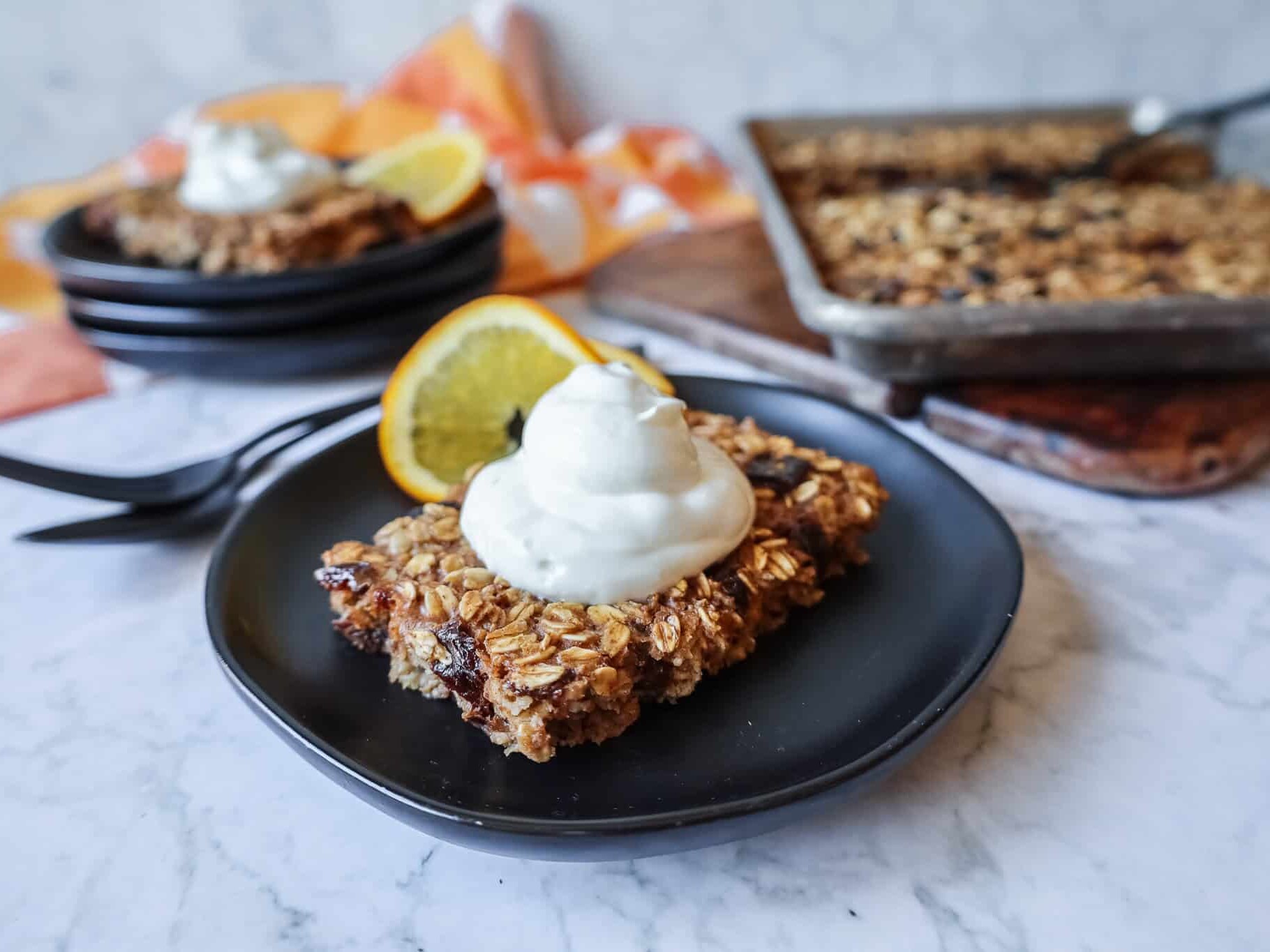 a slice of sheet pan baked oatmeal topped with yogurt and a sliced orange on a black plate