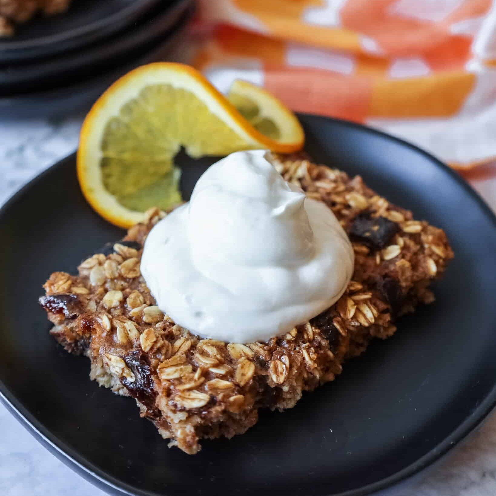 a slice of sheet pan baked oatmeal topped with yogurt and a sliced orange on a black plate